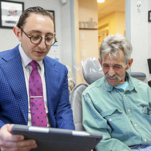 Doctor Ariel showing a tablet screen to a senior man in the dental chair